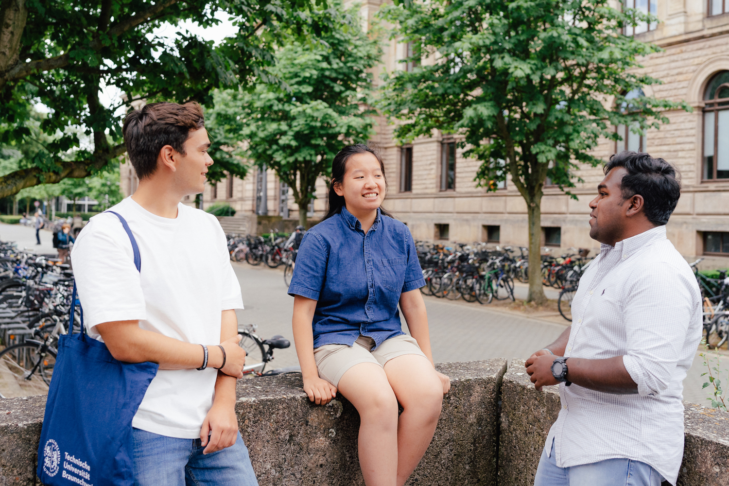 Three students stand in front of the old building of TU Braunschweig and chat animatedly with each other.